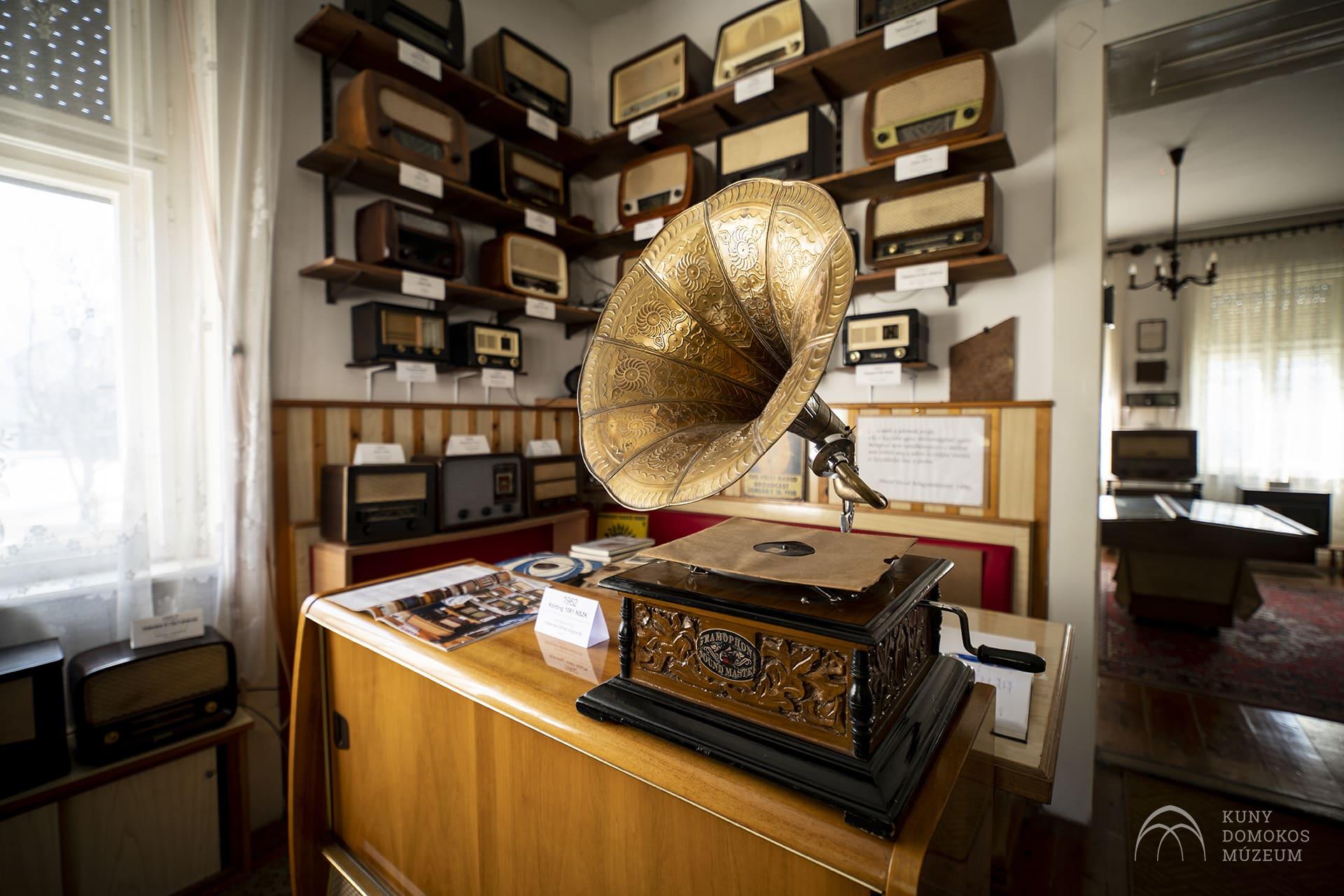Egy szoba, tele vintage rádiókkal és egy dekoratív antik gramofonnal egy fa vitrinen. A helyiségben fapolcok, különféle rádiómodellek, fehér falak, természetes fényt átengedő ablakokkal, a Kuny Domokos Múzeumot jelző tábla jelzi.