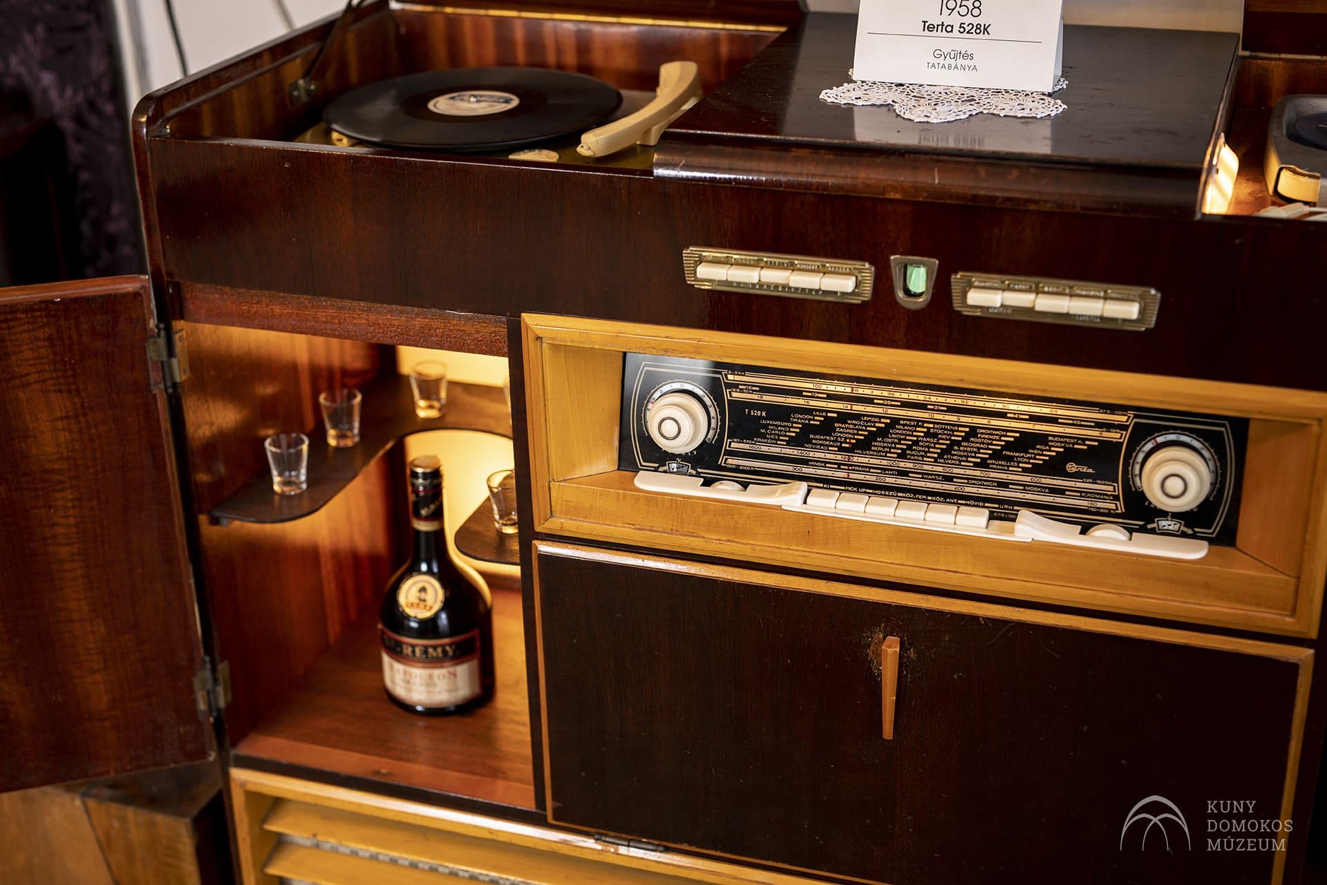 Egy vintage fa konzol lemezjátszóval bakelitlemezzel, régimódi rádiótunerrel és egy alsó szekrénnyel, egyetlen üveg itallal, két kis pohárral és egy füzettel. A tetején egy kis tábla található "1958" felirattal.