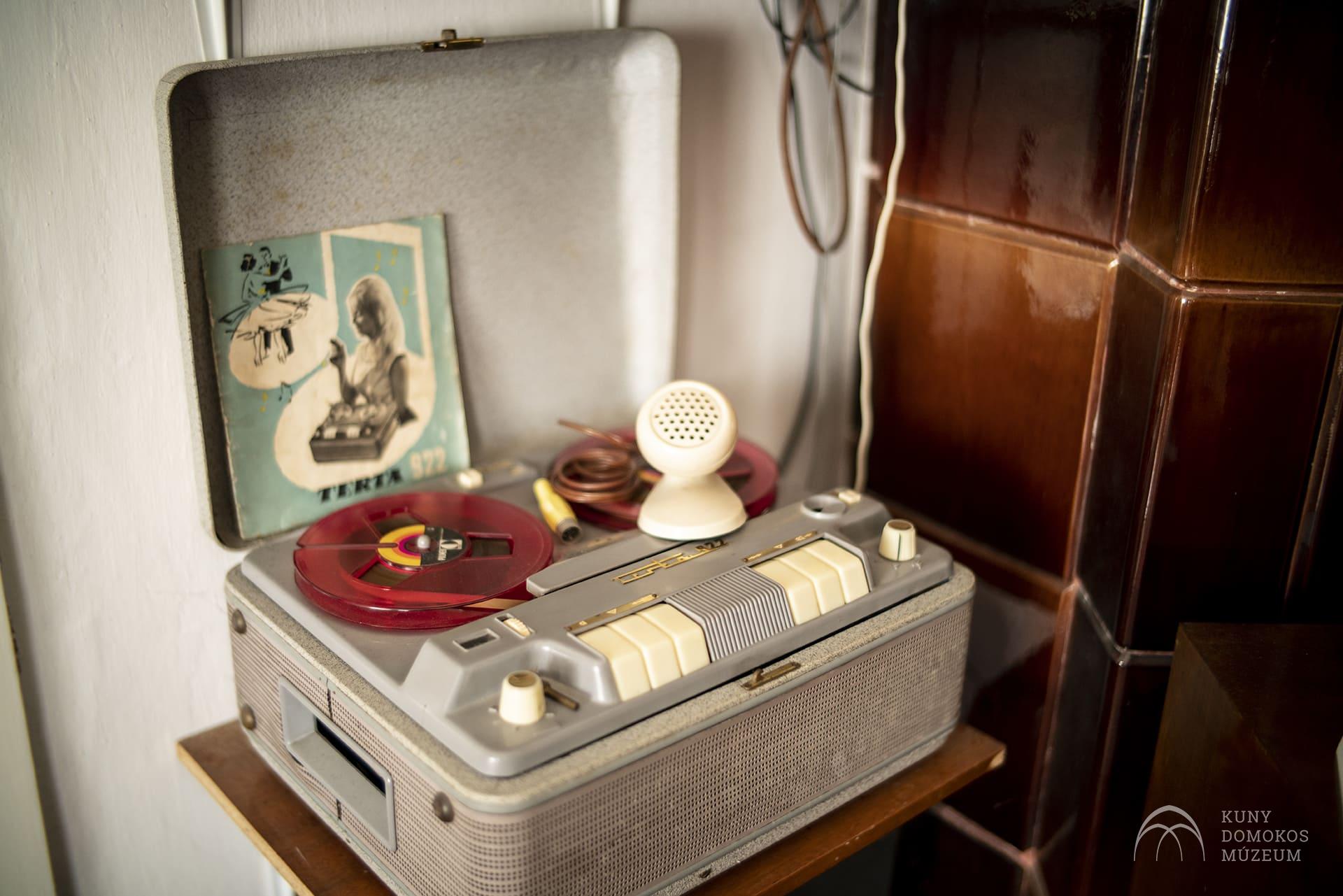 Egy vintage tekercses magnó ül nyitva egy fából készült asztalon. Két piros orsó van felszerelve rá, és egy kis fehér mikrofon pihen a tetején. Egy régi folyóirat van a gép mellett. A háttérben egy részben látható, kerámia csempével borított fa szekrény található.