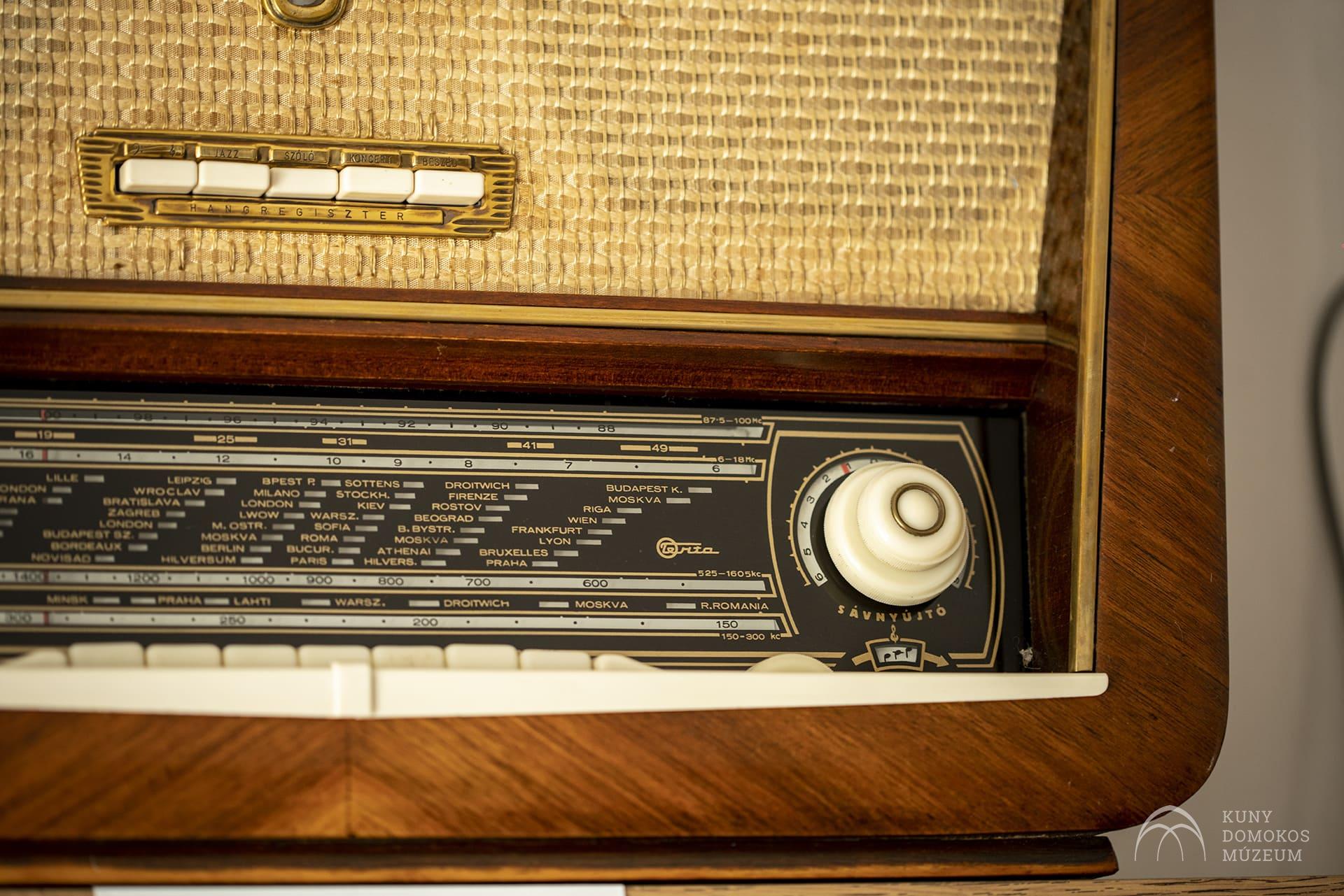 Közeli kép egy vintage rádióról szőtt szövet hangszóróburkolattal és fa furnér burkolattal. A vezérlőpulton nagy fehér hangológomb, városnevekkel és állomássávokkal ellátott frekvenciakijelző, a jobb alsó sarokban pedig a Kuny Domokos Múzeum logója található.