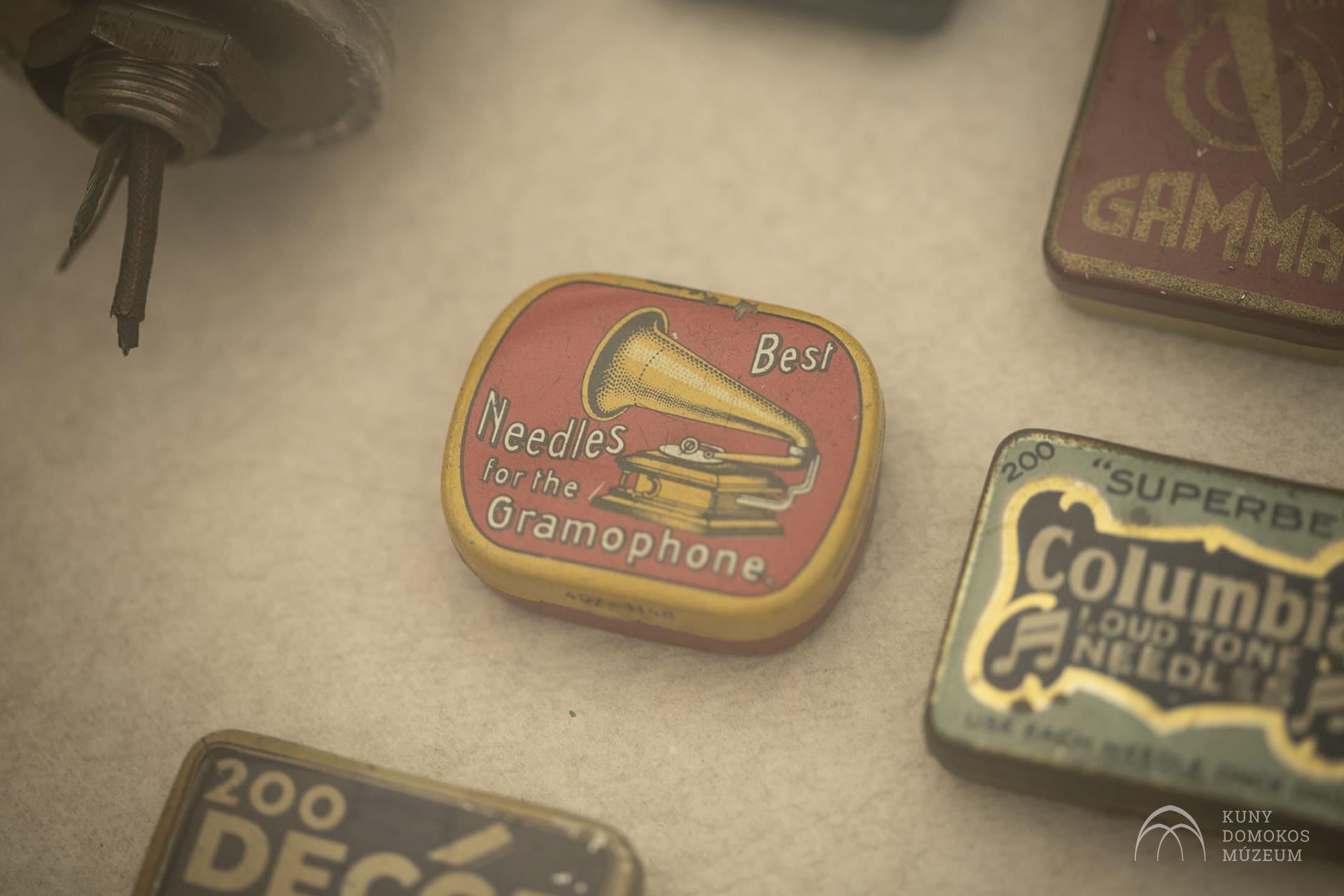 Egy kis, vintage bádogdoboz "Legjobb tűk a gramofonhoz" felirattal, a fedelén egy gramofon illusztrációja. A doboz egy olyan kollekció része, amely más, különböző feliratozású konzervdobozokat is tartalmaz. A háttér törtfehér felület.
