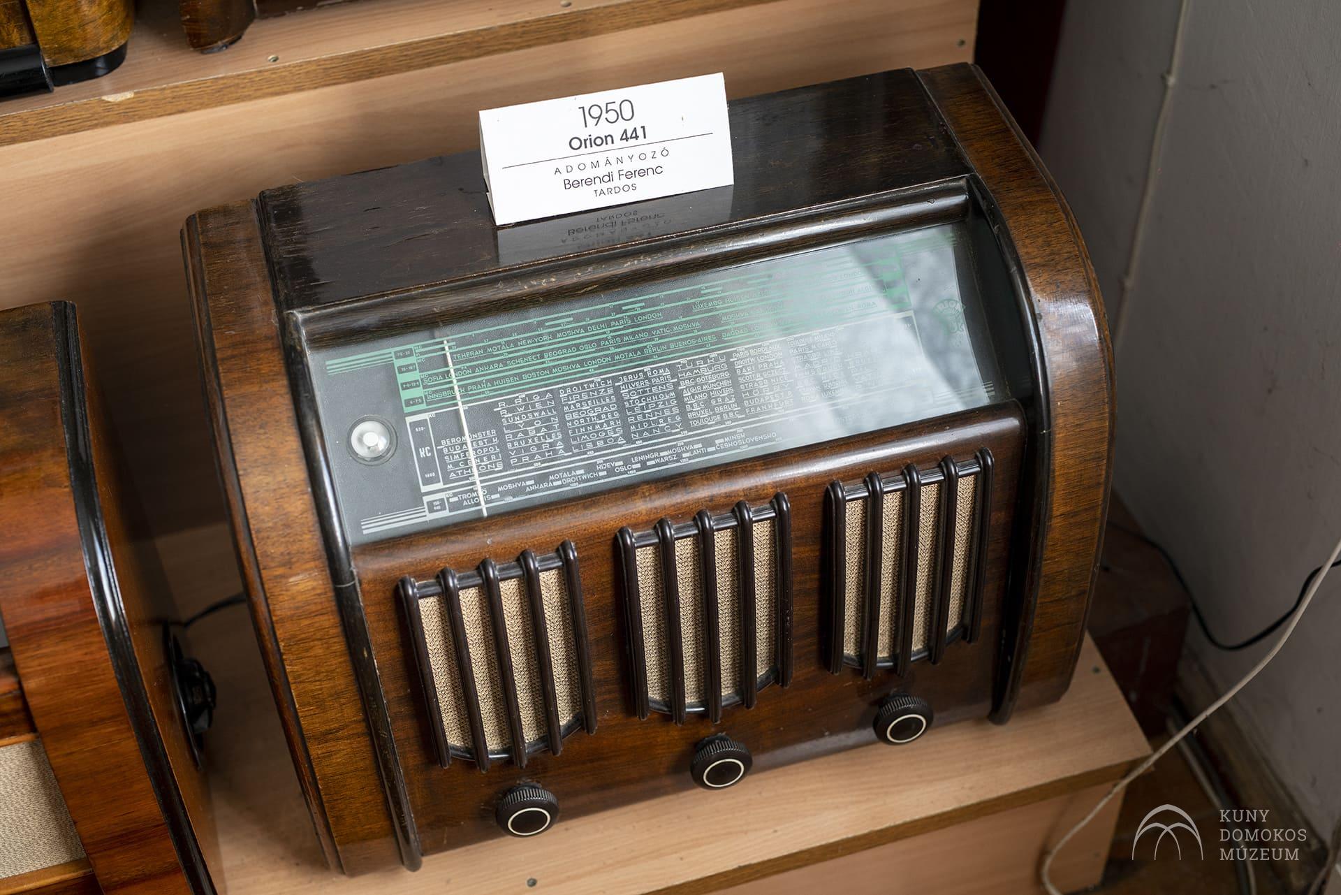 Egy régi rádió 1950-ből, "Orion 441" felirattal, egy fapolcon. A rádió fa külseje a frekvenciatárcsát bemutató üveglappal, valamint számos hangszórórács és vezérlőgomb az előlapon található. Felette egy kis tábla található a modell információival.