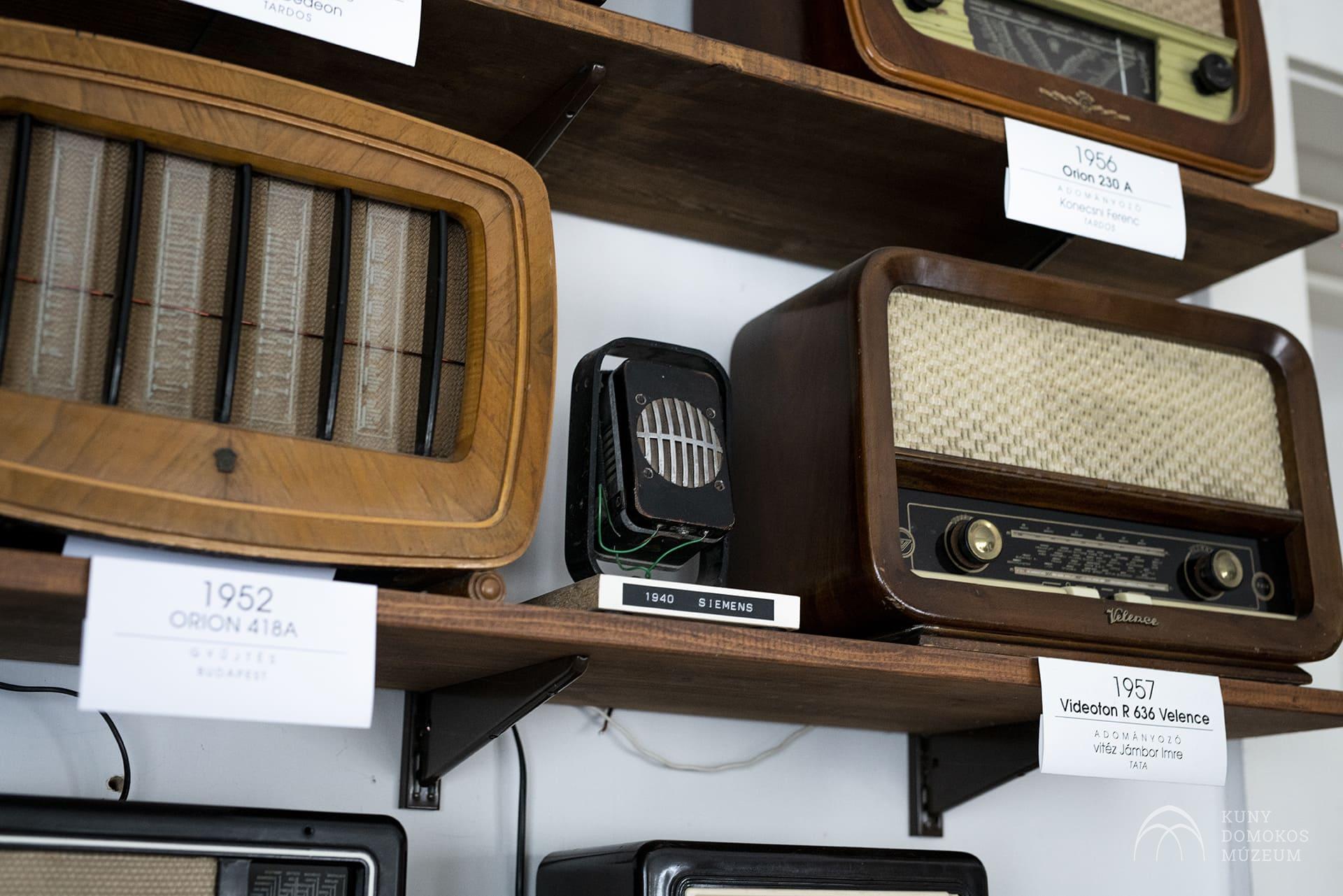 Régi rádiók gyűjteménye fapolcokon. A rádiók kialakítása és kora eltérő, a címkéken feltüntetik a megfelelő évszámot és modellt. A látható címkék közé tartozik az Orion 41BA (1952), a Siemens (1940) és a Videoton R-641 (1957).