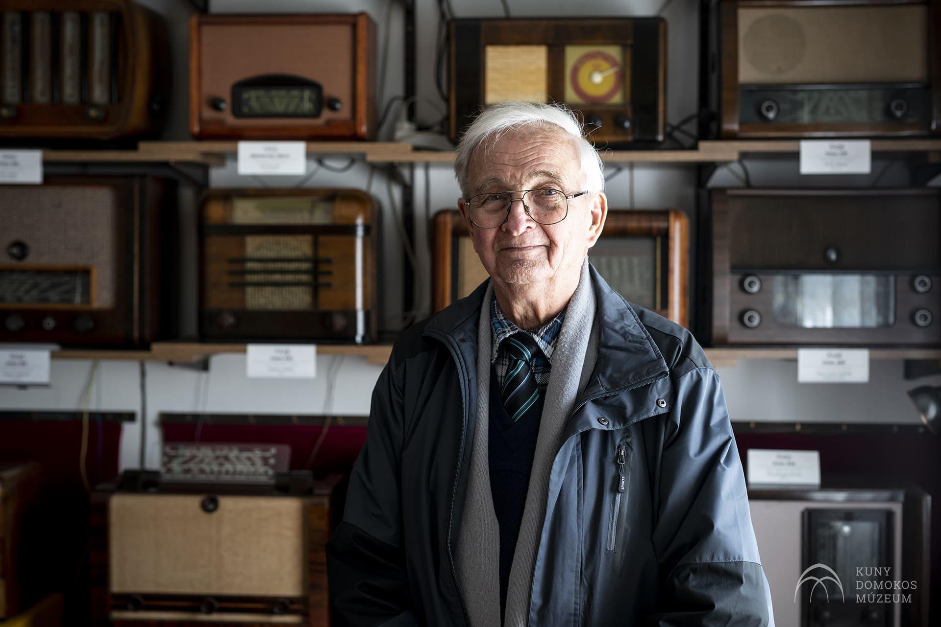 Egy idős, fehér hajú, szemüveges férfi áll a mögötte polcokon kihelyezett vintage rádiókkal teli szobában. Szürke kabátot, kék inget és csíkos nyakkendőt visel, és finoman mosolyog a kamerába. A sarokban múzeum logója látható.