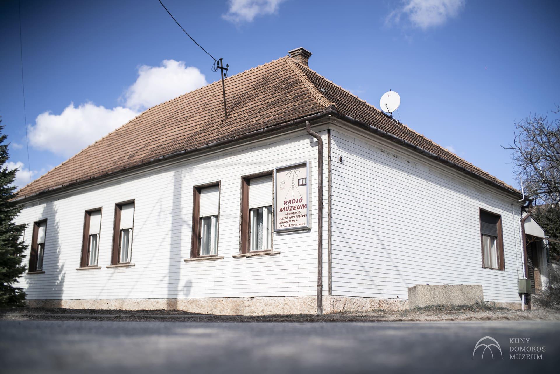 Egy kis fehér épület ferde vörös cseréptetővel, három oldali ablakokkal, az egyik ablaknál Rádió Múzeum feliratú tábla lóg. Tiszta, kék ég van, a háttérben néhány felhő, a jobb oldalon néhány csupasz fa látható.