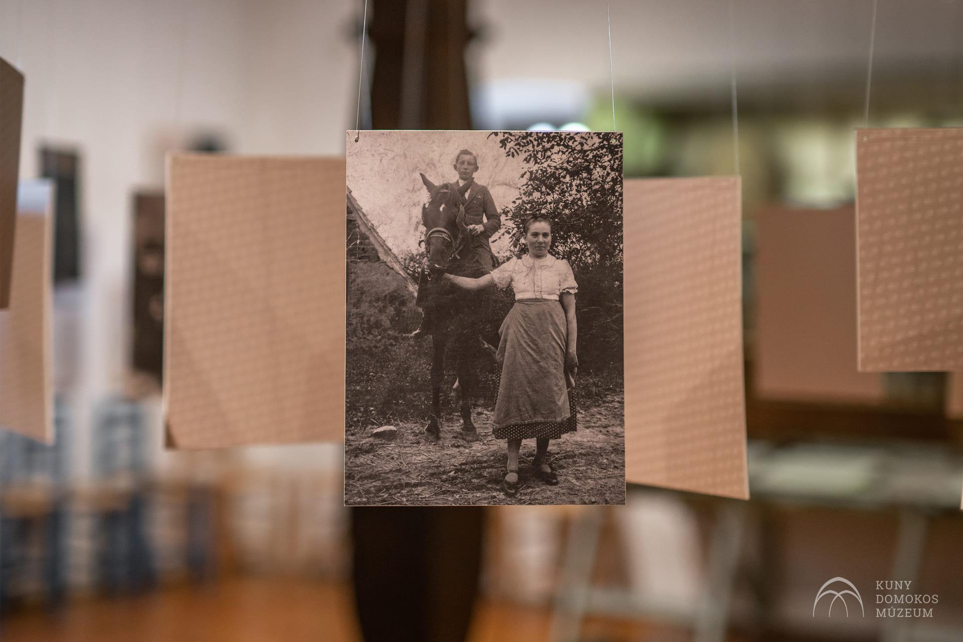 Egy ló mellett álló fiatal lány szüreti fekete-fehér fényképe látható egy kiállítás részeként. A lány térdig érő ruhába van öltözve, és közvetlenül a kamerába néz. A háttérben elmosódott, texturált papírok lógnak a fotó körül.