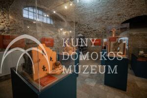 A téglafalakkal és boltíves ablakokkal díszített múzeumi kiállítóteremben több üveges vitrin látható, amelyek történelmi leleteket és másolatokat tartalmaznak. Az információs táblák és a részletes modellek kék állványokon helyezkednek el, amelyeket a mennyezetről reflektorok világítanak meg.