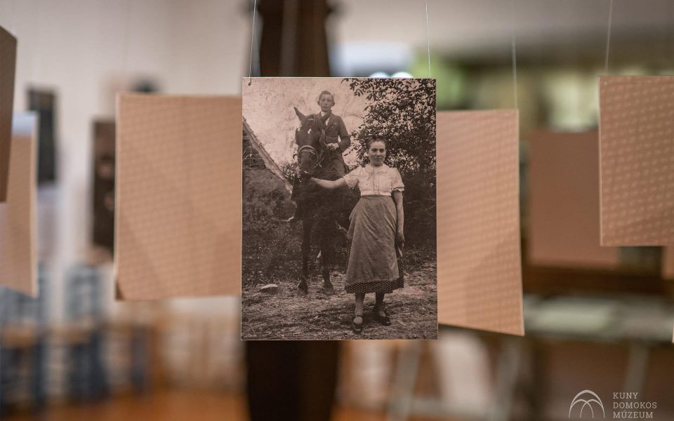 Egy ló mellett álló fiatal lány szüreti fekete-fehér fényképe látható egy kiállítás részeként. A lány térdig érő ruhába van öltözve, és közvetlenül a kamerába néz. A háttérben elmosódott, texturált papírok lógnak a fotó körül.